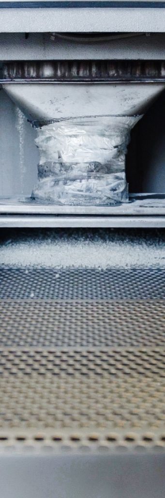 White plastic granules beneath a material feed outlet on a perforated metal sieve.