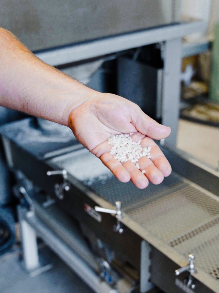 Eine offene Hand hält weißes Kunststoffgranulat vor einer industriellen Siebanlage.